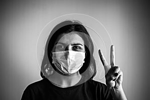 Portrait of a young woman wearing medical sterile mask, showing victory sign with her hand. Dramatic black and white closeup of a