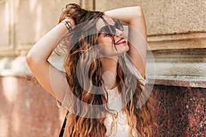 Portrait of young woman wearing sunglasses with long hair smiling and looking up. Young female student posing in the city street