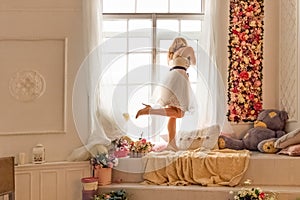 Portrait of a young woman in a white dress against the background of a closed large window, looking out into the street through