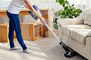 Portrait of young woman in white shirt and jeans cleaning carpet under sofa with vacuum cleaner in living room, copy space.