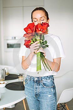 Portrait of young woman in white t-shirt with bouquet of red flowers in hands near window in bright kitchen at home