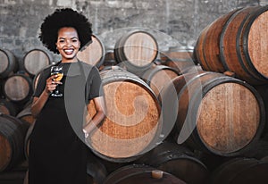 Portrait of a young woman winemaker standing with a glass with wooden barrel of red wine in a winery cellar or