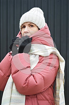 Portrait of a young woman in a winter down jacket, knitted hat, gloves and a light scarf on a dark background
