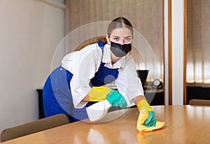 Portrait of a young woman worker in a protective mask, washing an office desk