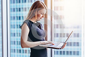 Portrait of young woman working holding laptop standing against panoramic window with city view