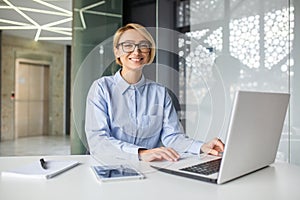 Portrait of a young woman working in the office using a laptop, looking and smiling at the camera while sitting at a