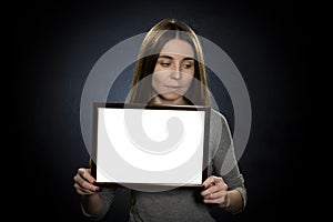 Portrait of a Young woman 25-30 years old holding a white text frame on a dark background, shyly averting her eyes to the side photo
