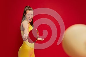 Portrait of young woman in yellow sportswear posing, playing table tennis isolated over red background. Hobby