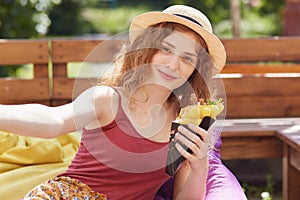 Portrait of young women sitting in open air on frameless chair, eating ice cream and taking selfie photo in summer park, relaxing