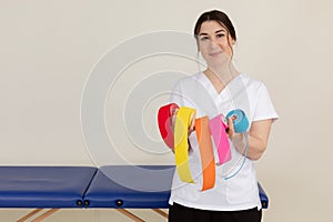 Portrait of young wonderful masseuse woman wearing white uniform, showing colourful kinesio tapes rolls near blue couch.