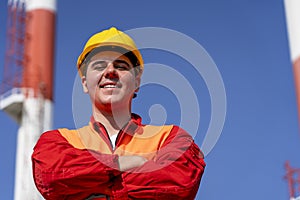 Portrait of Young Worker in Red Coveralls and Yellow Hardhat Against Power Plant Chimneys and Blue Sky