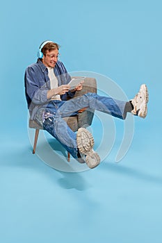 Portrait of youngemotional man in headphones sitting on chair and playing games on tablet over blue studio background