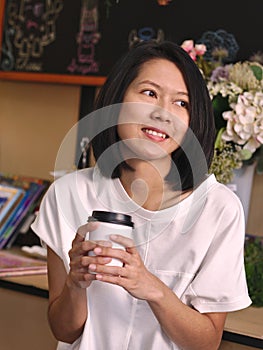 Portraits of Asian woman holding a cup of coffee by two hands looking to her left hand in cozy coffee shop.