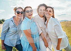 Portraits of four cheerful smiling women in sunglasses embracing and looking at camera during outdoor walking with bright sunny