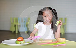 Portraits of happy Asian child girl slicing cucumber vegetable on chopping board at play room. Kid play chef cooking