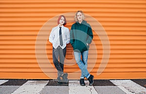 Portraits of two caucasian teen friends boy and girl posing for photo while they standing near the orange wall background.