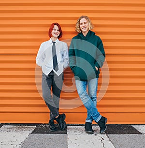 Portraits of two smiling caucasian teen friends boy and girl posing for photo while they standing near the orange wall background