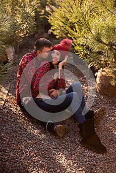 Portraits of young couple in forest, wearing red checkered shirts sitting among Christmas tree