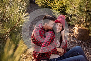 Portraits of young couple in forest, wearing red checkered shirts sitting among Christmas tree