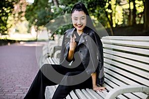 Portrat of young pretty laughing Asian woman in fashionable black pants and leather jacket, sitting on the bench green