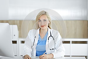 Portrate of woman-doctor at work while sitting at the desk in clinic. Blonde cheerful physician filling up medical form