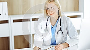 Portrate of woman-doctor at work while sitting at the desk in clinic. Blonde cheerful physician ready to help patients