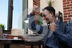 Portratit of startup marketing specialist with glasses doing celebration hand gesture sitting at desk