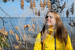 Portret of happy smiling young woman with her eyes closed enjoys the sunshine and fresh air. She stands in against lake
