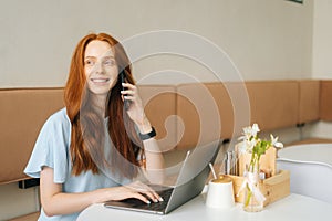 Portrtait of happy young woman talking on smartphone and using laptop sitting at table by window in cozy light cafe