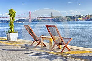 Portugal Travel Ideas. Line of Wooden Cahris Placed in Front of 25 April Bridge in Lisbon At Tagus River Shoreline in Lisbon in