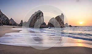 Portugal Ursa Beach at coast of Atlantic Ocean. Sea stack rocks in sunset lit. Foamy waves rolling to sandy coastline