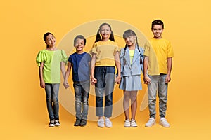 Positive adorable diverse children holding hands and smiling on yellow