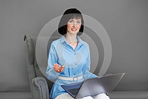 Positive adult business woman mentor teacher psychologist sits on armchair consulates talk in online seminar on laptop