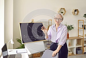 Positive adult gray haired woman teacher stands next to table with laptop and blackboard