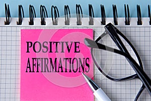 POSITIVE AFFIRMATIONS - words on a pink piece of paper on a white notebook with glasses and pen