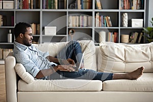 Positive African American man lying on couch, using laptop