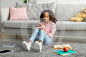 Positive african american teen girl doing homework, making notes or writing essay, sitting in living room interior