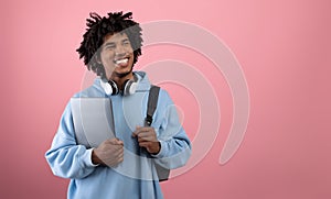 Positive African American teen student with backpack, tablet pc and headphones studying online on pink background