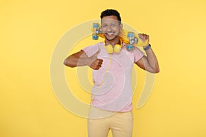 Positive african american young man wearing headphones holding skateboard over yellow background. Show thumb up.