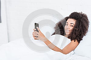 Positive african american young woman using smartphone in bed during morning time