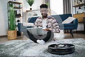 Positive african guy using robotic vacuum cleaner for chores