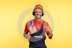 Positive afro-american man in blue overalls red t-shirt and cap holding usa flag and showing thumbs up, happy satisfied with life