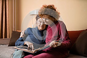 Positive aged ladies looking album photos sitting sofa at home, cheerful friends. Senior woman and her mature nurse watching photo