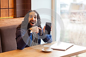 Positive amazing handsome young adult man freelancer in casual style sitting in cafe, pointing finger to empty screen phone,
