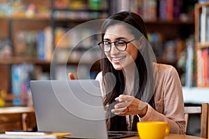 Positive arab woman having video conference on laptop, sitting at city cafe, talking and gesturing to computer webcamera