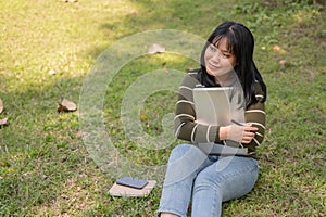 A positive Asian female college student is sitting on the grass in a campus park with her laptop