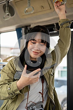 A positive Asian female solo traveler using her smartphone while standing in a public bus