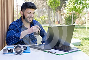 Positive asian guy with beard working at laptop outdoors.