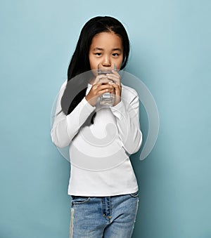 Positive asian Korean girl in jeans and white long sleeve standing and drinking pure water from glass over blue