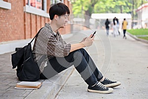 A positive Asian male college student sits on the street near the campus building using his phone
